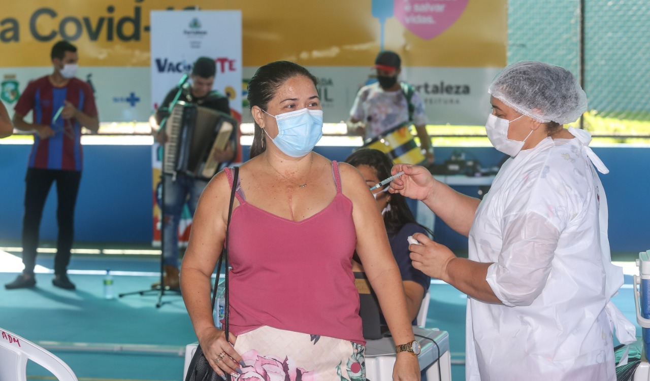 mulher de camisa rosa e máscara recebe a vacina de uma enfermeira em um ponto de vacinação. Atrás ocorre apresentação musical com três músicos