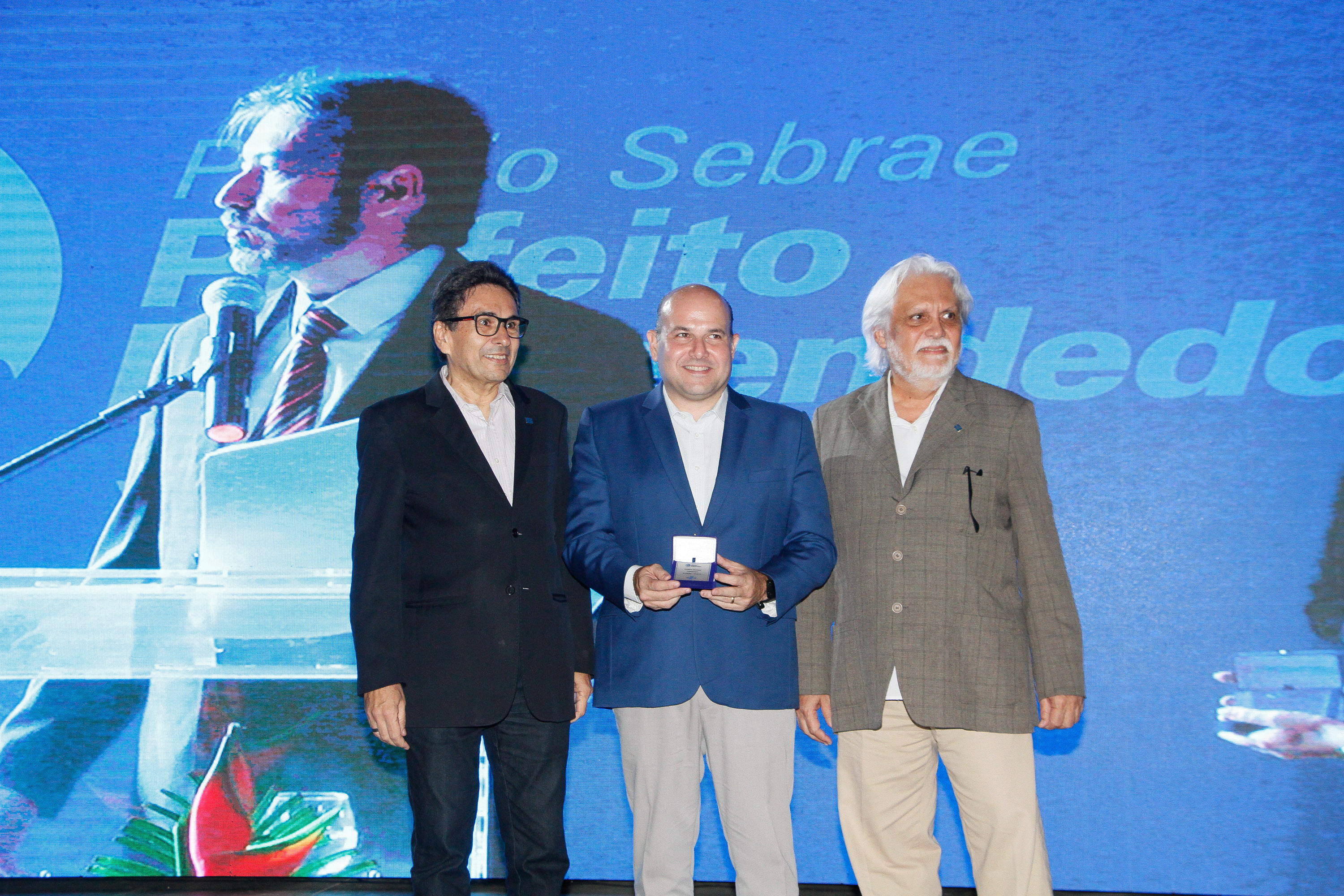 Prefeito no palco, em meio a dois senhores, segura o prêmio recebido