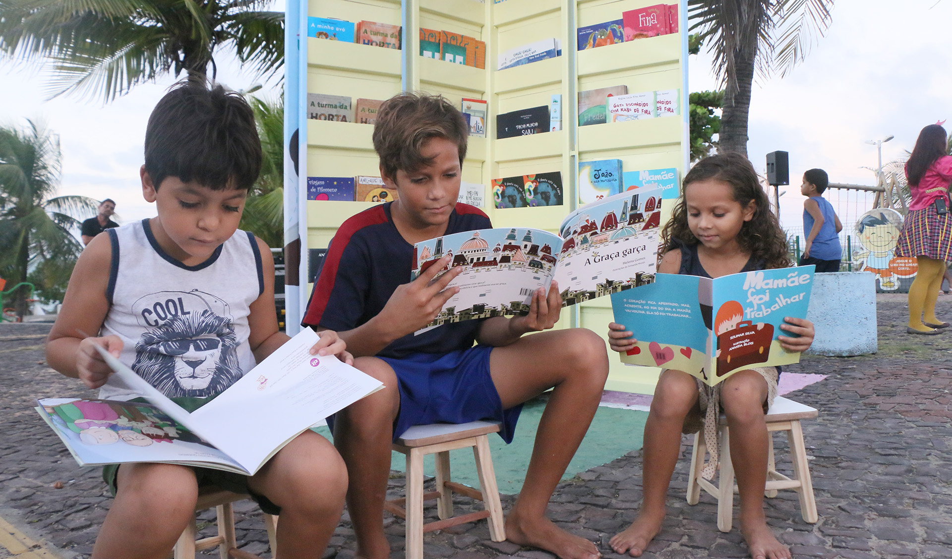 três crianças leem livros sentadas em banquinhos na praça