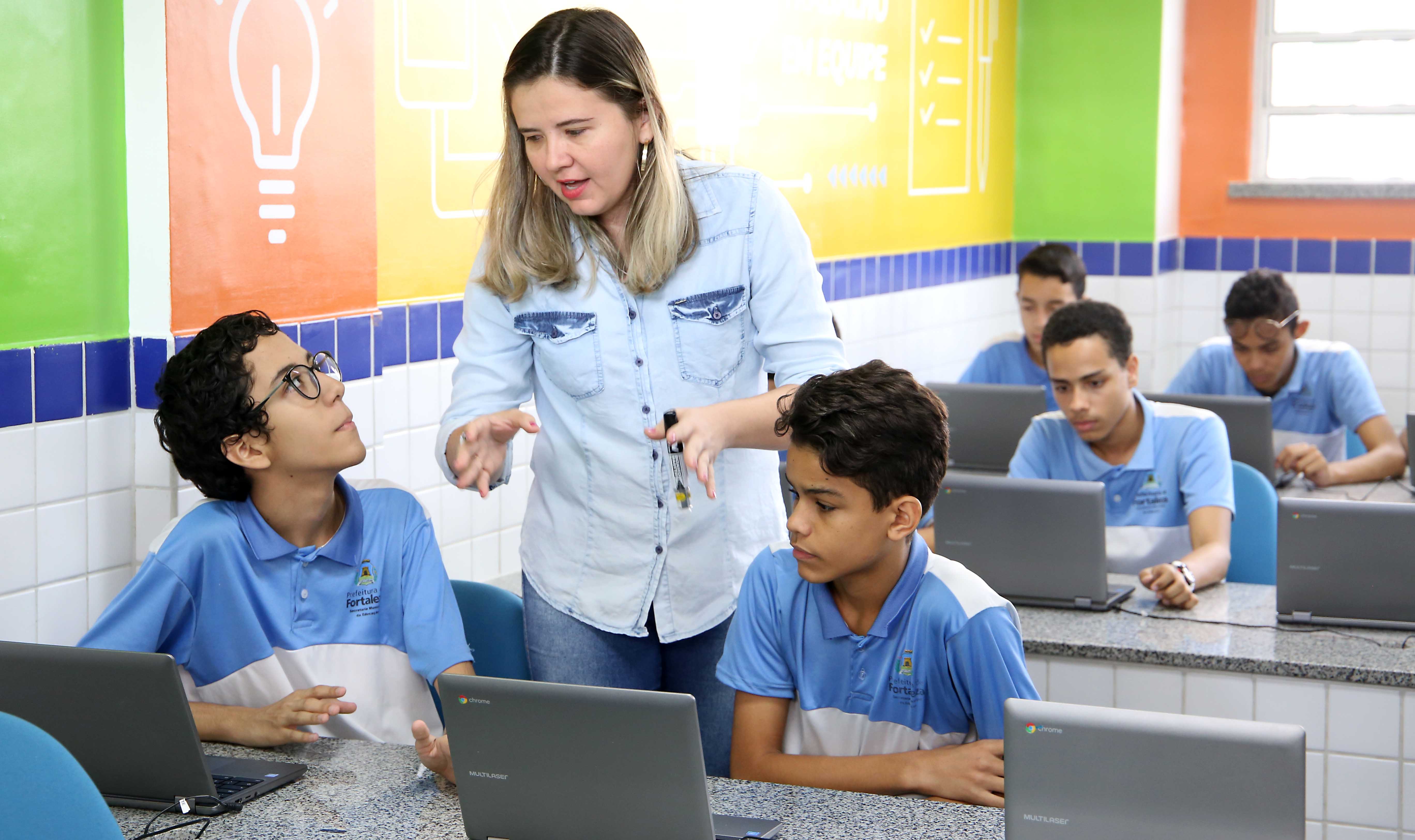 professora em pé falando entre dois alunos sentado em frente a computadores