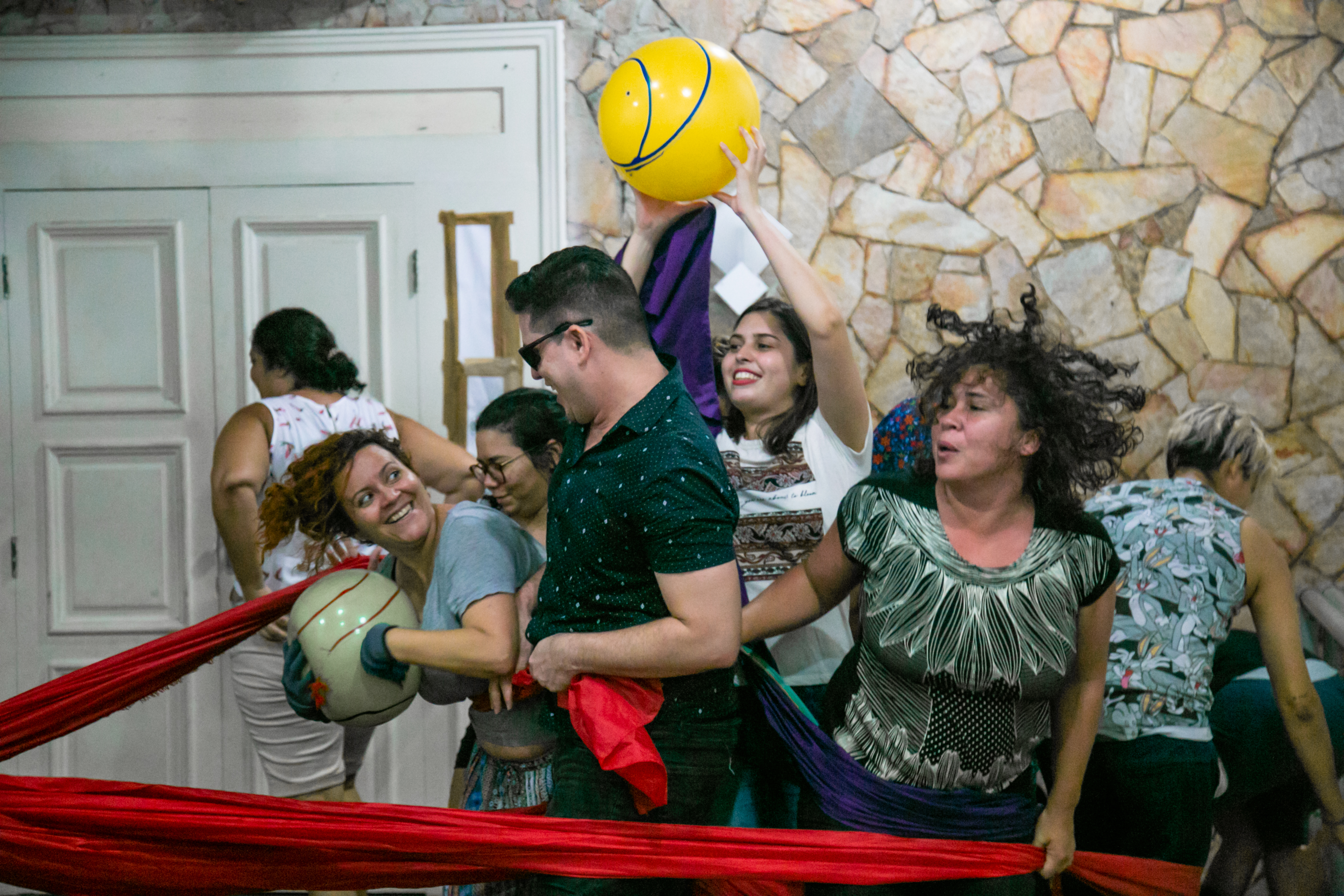 Numa dinâmica teatral, pessoas enlaçadas por um grande lenço vermelho brincam com bolas de látex.