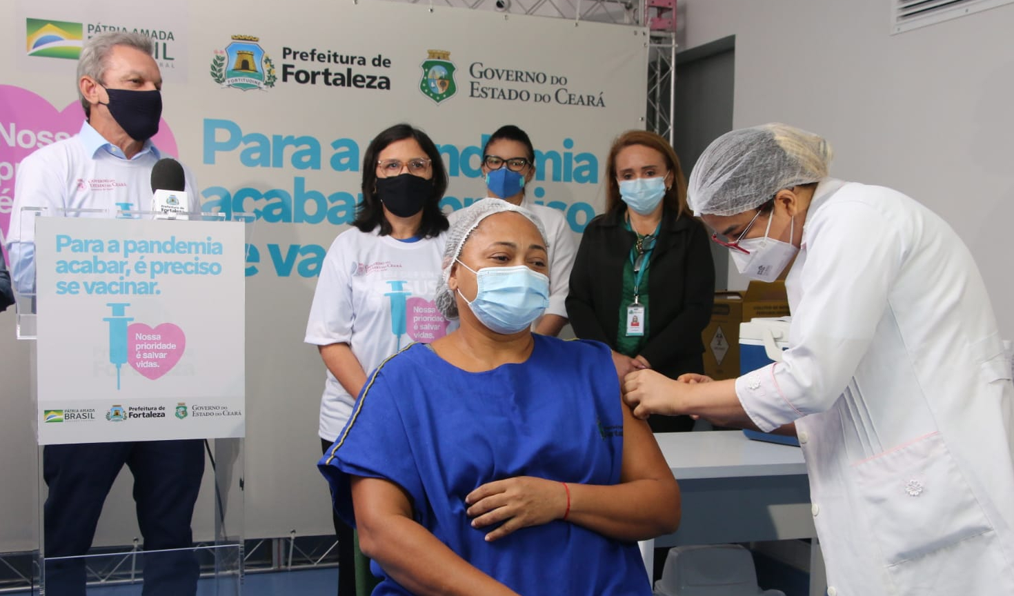 José Sarto em pé ao lado de Ana Estela Leite e outras pessoas assistindo enfermeira sentada à frente ser vacinada