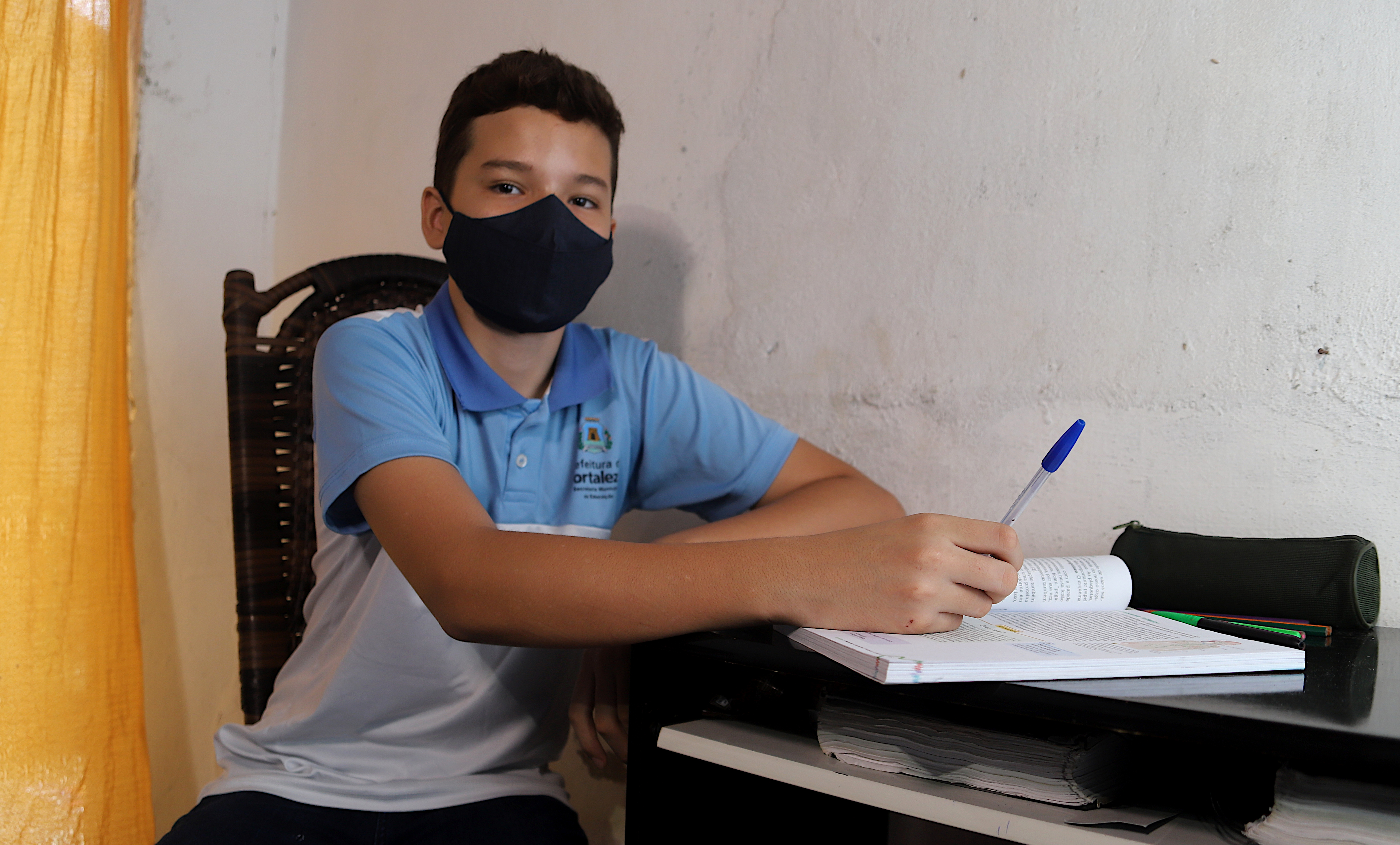 garoto vestindo farda escola da rede municipal e usando máscara sentado à mesa com livro e segurando caneta