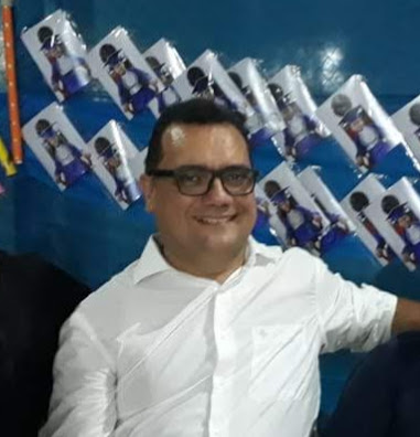 professor sorrindo para a foto com mural de fotos de alunos em cadeiras de formatura ao fundo