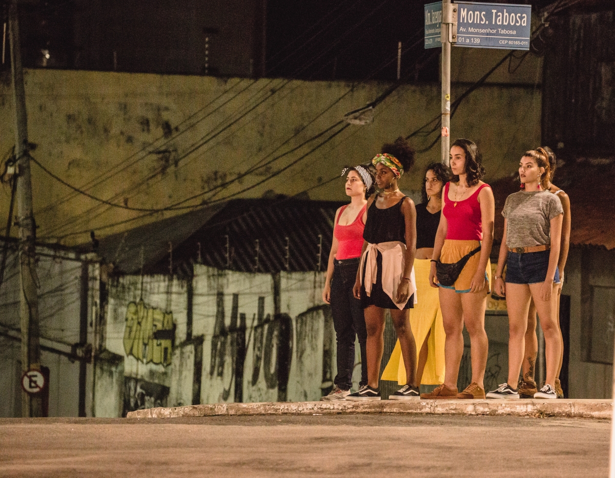 Grupo de mulheres com roupas coloridas paradas em esquina de rua em que se vê a placa com 