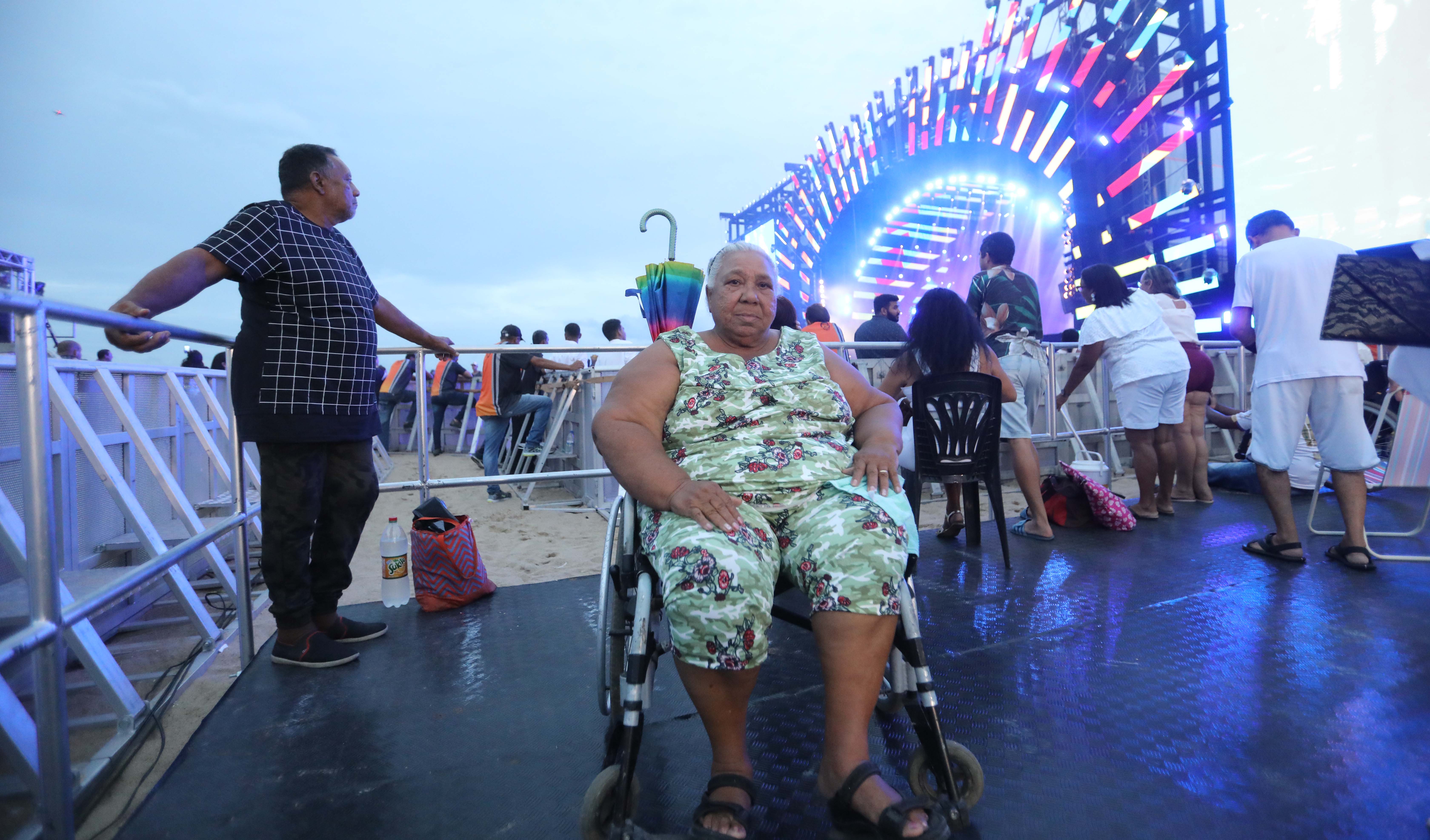 senhora sentada em cadeira de rodas posando para a foto com palco e pessoas ao fundo