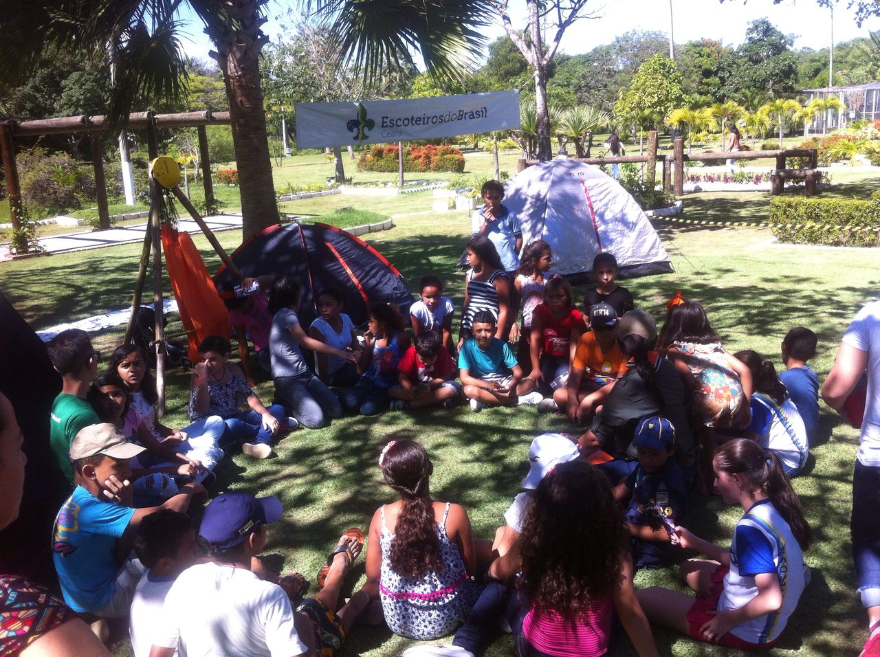 Crianças sentadas num gramado verde recebem instruções das atividades no Grupo de Escoteiros Sargento Prata