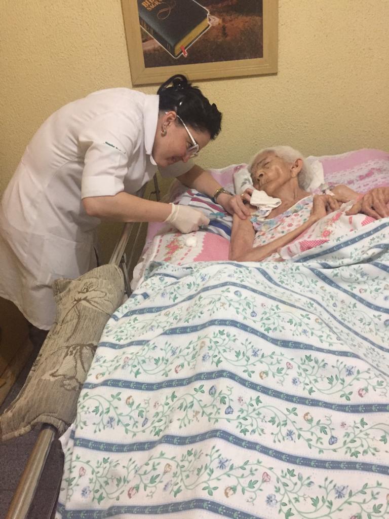Mulher aplica vacina em idosa deitada em cama